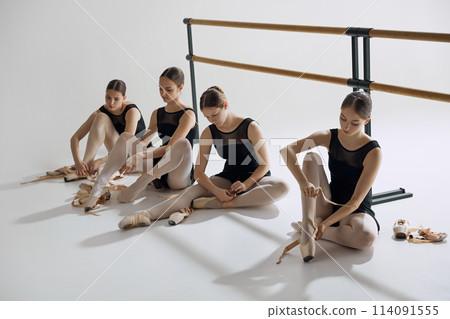 Behind the scenes of ballet. Four elegant teen girls, baller dancers sitting on floor, getting ready for dance, adjusting pointe shoes. 114091555