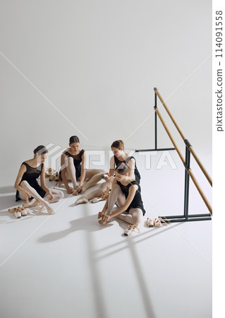 Behind the scenes of ballet. Four elegant teen girls, baller dancers sitting on floor, getting ready for dance, adjusting pointe shoes. 114091558
