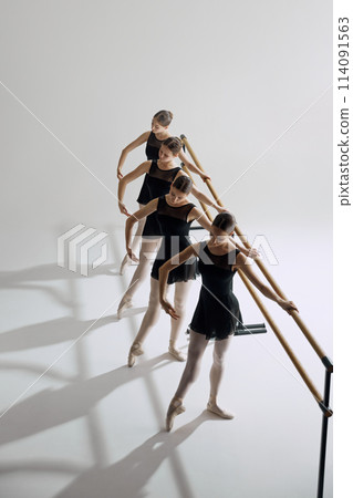 Four elegant girls, ballet dancers standing at barre and warming up, practicing against grey studio background. Focus on discipline 114091563