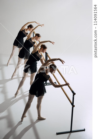 Symmetry and grace in ballet. Four flexible girls, ballet dancers standing at barre and training against grey studio background Symmetry and grace in ballet. Four flexible girls, ballet dancers standing at barre and training against grey studio background 114091564
