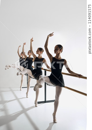 Four girls, ballet dancers in studio standing at barre, reaching out gracefully with one arm, practicing against grey background. 114091575