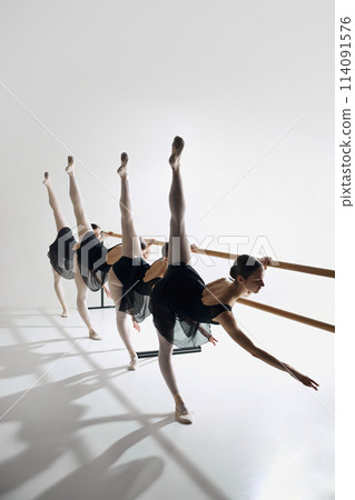 Four ballet dancers line up at barre, performing identical pose with outstretched leg, training against grey studio background 114091576