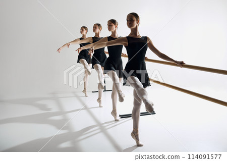Four elegant girls, ballet dancers standing at barre and warming up, practicing against grey studio background. Focus on discipline 114091577