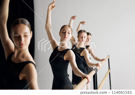 Growing skills before performance. Teen beautiful girls, ballet dancers standing at barre and practicing with concentration against grey studio background 114091583