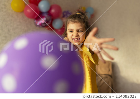 Smiling real girl 7 years old in yellow dress catches violet balloon against festive backdrop in holiday at home 114092022