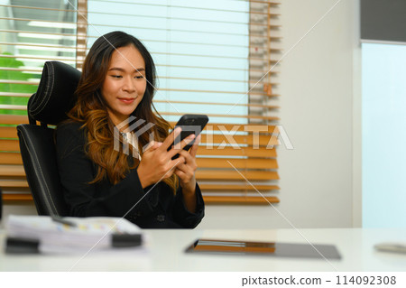 Attractive businesswoman texting messages on smartphone sitting in office Attractive businesswoman texting messages on smartphone sitting in office 114092308