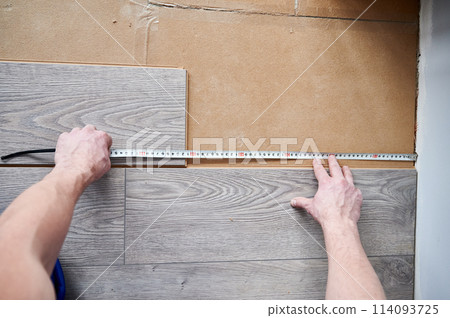 Close up of male construction worker using tape measure ruler while measuring distance from wall to laminate board. Man installing laminate flooring in apartment under renovation. 114093725