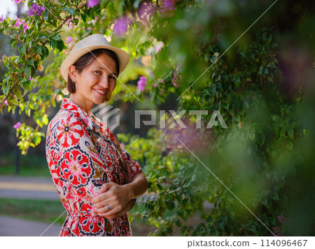 young asian woman stands in sunny green park, admiring blooming bougainvillea bush. The flower's bright colors are beautiful contrast to park's lush greenery. 114096467