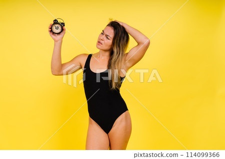 Female holding alarm clock, isolated on yellow. Young model in black swimsuit. 114099366