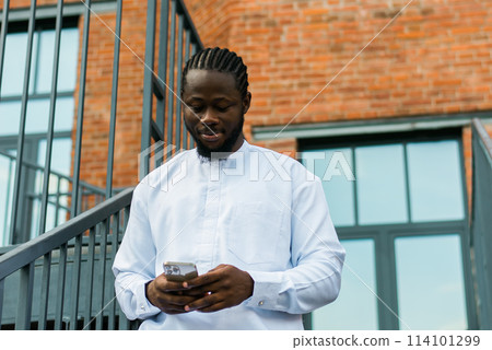 African american man in stylish ethnic dashiki wear viewing photos in social networks on mobile enjoying free time outdoors. Dark-skinned man watching funny video on smartphone and social networks African american man in stylish ethnic dashiki wear viewing photos in social networks on mobile enjoying free time outdoors. Dark-skinned man watching funny video on smartphone and social networks 114101299