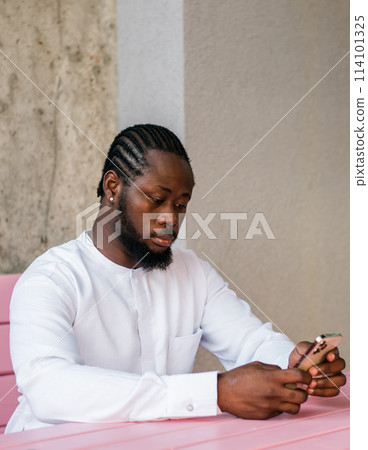 African American hipster guy in cultural and ethnic clothing dashiki chatting on smartphone outdoors, texting with friends on social networks. Millennial generation and Internet blog concept 114101325
