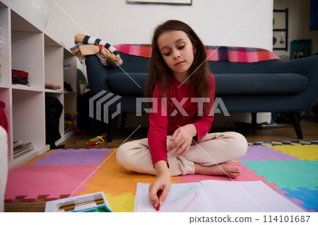 Little child girl with long hair, sitting on the puzzle carpet at home, doing homework, studying, drawing picture with colorful pencils. People, kids education and entertainment. Art and creativity 114101687