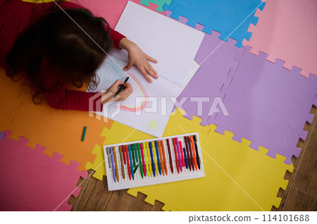 Little girl smiling drawing picture, lying on her belly on a multicolored puzzle carpet. People. Education. Back to school concept Little girl smiling drawing picture, lying on her belly on a multicolored puzzle carpet. People. Education. Back to school concept 114101688