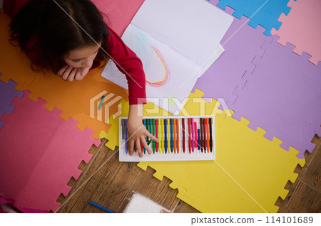 View from above of a little girl taking out colorful pencil from pencil case, drawing beautiful cloud with rainbow, lying on a multi colored puzzle carpet in cozy home interior. Kids education 114101689