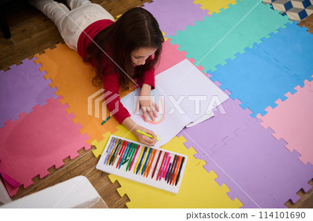 Top view of a talented little child, school girl taking out colorful pencil from pencil case, drawing beautiful cloud with rainbow, lying on a multi colored puzzle carpet in cozy home interior 114101690