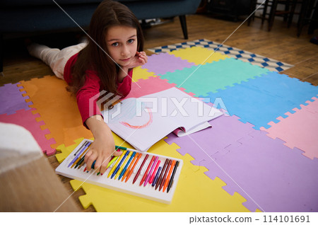 Little girl smiling looking at camera while taking a colorful pencil out of a box with pastel crayons, lying on her belly on a multicolored puzzle carpet. People. Education. Back to school concept 114101691
