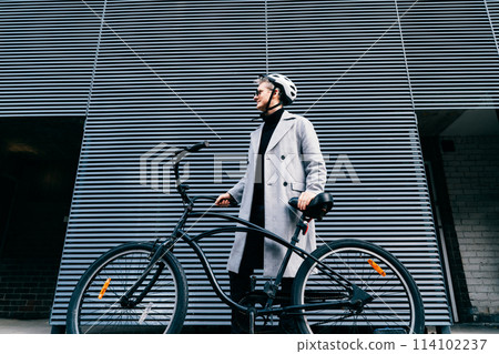 Stylish male in coat, sunglasses and protective helmet standing with retro bicycle near gray urban wall. Neutral carbon footprint transportation. Green eco friendly mobility sustainable transport. 114102237