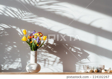 Scandinavian home interior with colorful bouquet of various flowers in ceramic vase standing on wooden cabinet under sunlight and shadows on white gray wall. Minimalist design of home decor. 114102267