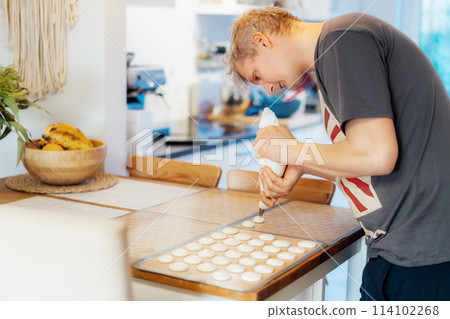 Concentrated young man placing batter on silicone baking sheet to make French macaroons dessert in light modern kitchen at home. Preparing steps for baking cookies. Culinary hobby. Selective focus Concentrated young man placing batter on silicone baking sheet to make French macaroons dessert in light modern kitchen at home. Preparing steps for baking cookies. Culinary hobby. Selective focus 114102268