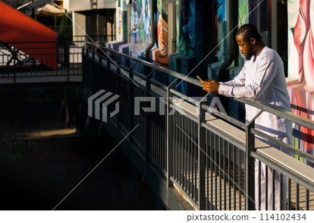 African american man in stylish ethnic dashiki wear viewing photos in social networks on mobile enjoying free time outdoors. Dark-skinned man watching funny video on smartphone and social networks 114102434