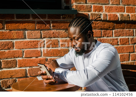 African american man in stylish ethnic dashiki wear viewing photos in social networks on mobile enjoying free time outdoors. Dark-skinned man watching funny video on smartphone and social networks African american man in stylish ethnic dashiki wear viewing photos in social networks on mobile enjoying free time outdoors. Dark-skinned man watching funny video on smartphone and social networks 114102456