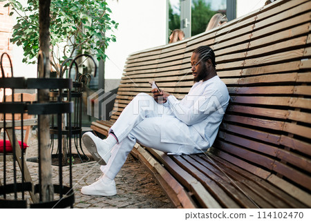 African american man in stylish ethnic dashiki wear viewing photos in social networks on mobile enjoying free time outdoors. Dark-skinned man watching funny video on smartphone and social networks 114102470