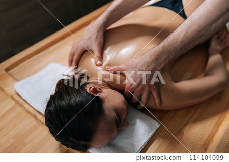 Top view of relaxed woman with closed eyes during professional massage in wellness center. Closeup of strong masseur male hands massage back. Pleasant atmosphere of spa salon with dark interior. 114104099