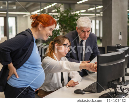 An elderly man, a Caucasian woman and a pregnant woman are discussing work issues at the computer.  114105482