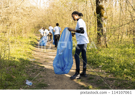 African american girl collecting garbage in a disposal blue bag, supporting nature preservation and helping protect the environment. Proud teenager volunteering to pick up trash. African american girl collecting garbage in a disposal blue bag, supporting nature preservation and helping protect the environment. Proud teenager volunteering to pick up trash. 114107061
