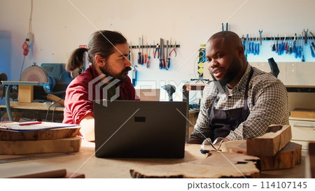 Cabinetmakers doing brainstorming while using CAD software on laptop, discussing wood designs. Carpenters in assembly shop discussing, using program on notebook to plan furniture assembling, camera B Cabinetmakers doing brainstorming while using CAD software on laptop, discussing wood designs. Carpenters in assembly shop discussing, using program on notebook to plan furniture assembling, camera B 114107145