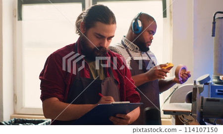Craftsperson drawing blueprints on notepad in studio with BIPOC coworker in background solving tasks. Manufacturer looking at technical schematics to execute woodworking projects, camera A Craftsperson drawing blueprints on notepad in studio with BIPOC coworker in background solving tasks. Manufacturer looking at technical schematics to execute woodworking projects, camera A 114107150