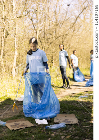 Community service volunteer reduces waste and cleans up forest setting, collecting garbage in blue trash bags. Young kid helps environment conservation by disposing of plastic waste. 114107169