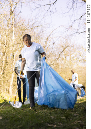 African american volunteer tidying the woods area of garbage and plastic bottles, collecting trash with claw and bags. Young woman doing voluntary work to conserve natural habitat. 114107172