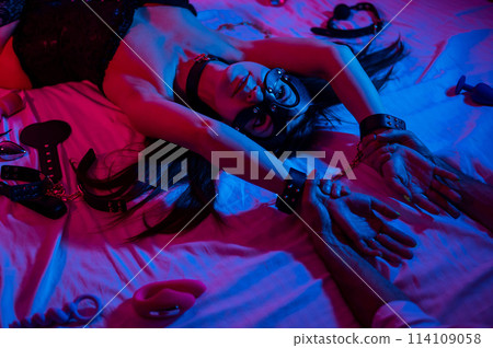 A woman in a leather mask and handcuffs lies on a bed in a blue-red light. Sex toys collection. A woman in a leather mask and handcuffs lies on a bed in a blue-red light. Sex toys collection. 114109058