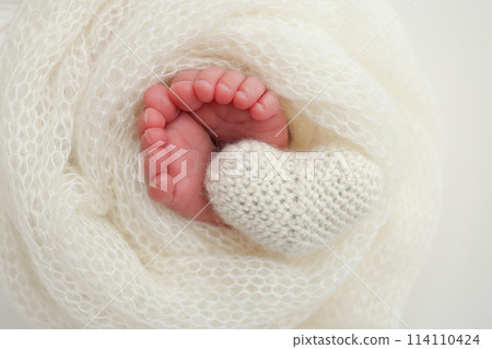 The tiny foot of a newborn baby. Soft feet of a new born in a white wool blanket. Close up of toes, heels and feet of a newborn. Knitted white heart in the legs of a baby. Macro photography The tiny foot of a newborn baby. Soft feet of a new born in a white wool blanket. Close up of toes, heels and feet of a newborn. Knitted white heart in the legs of a baby. Macro photography 114110424