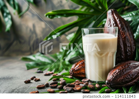 Fresh cacao drink in a transparent glass, surrounded by whole and split cacao pods and scattered beans, with lush green leaves in the background 114111843