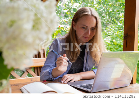 Female student has online lesson education outdoor in garden wooden alcove. Blonde woman sitting outside work on laptop having video call Female student has online lesson education outdoor in garden wooden alcove. Blonde woman sitting outside work on laptop having video call 114113292