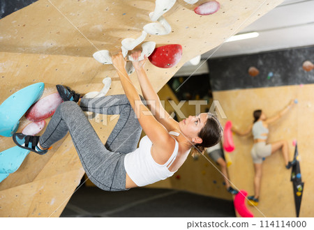 Young woman climbs steep artificial wall in sports complex and trains endurance 114114000