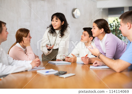 Young Chinese girl student discussing learning assignment with coursemates 114114069