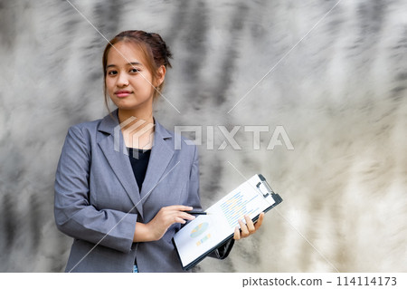 Young happy Asian businesswoman in a suit holding a pen and graph file clipboard, standing confident against the black and white background 114114173