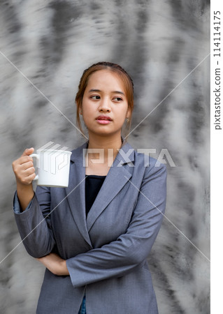 Young happy Asian businesswoman in a suit holding a mug of coffee, standing confident against the black and white background 114114175