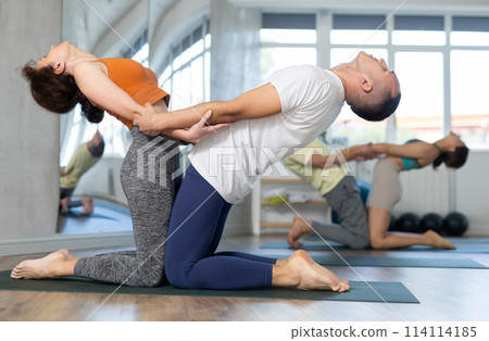 Lovers of active lifestyle perform pair yoga supported backbend on knee in gym Lovers of active lifestyle perform pair yoga supported backbend on knee in gym 114114185