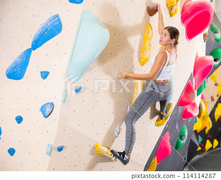Young woman climbs steep artificial wall in sports complex and trains endurance 114114287