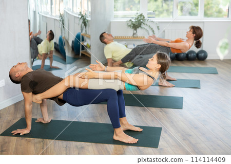 Young men and women maintaining healthy lifestyle practicing partner yoga at group lesson, training balance pose at gym 114114409