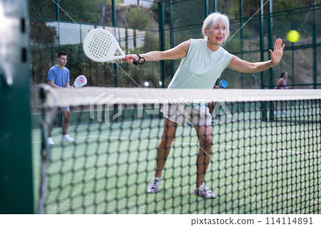 Aged woman playing friendly paddleball match on outdoor court Aged woman playing friendly paddleball match on outdoor court 114114891