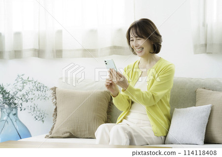 一位微笑的中年婦女在客廳操作智慧型手機 一位微笑的中年婦女在客廳操作智慧型手機 114118408