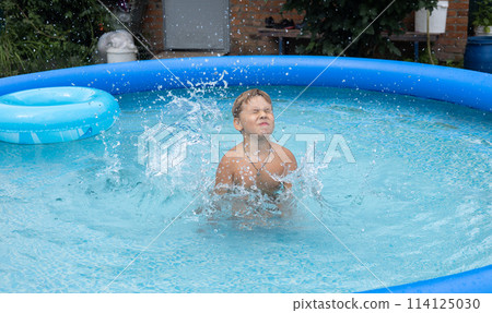 In the summer, with his eyes closed, a child jumps into the water in an inflatable pool and raises a lot of splashes. A child enjoys swimming in an inflatable pool in the yard on a hot summer day. 114125030