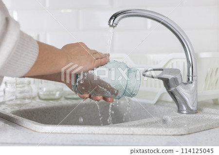 In the kitchen sink, a woman under the tap washes a glass jar to be sent for recycling. A woman sorts garbage to be sent for recycling and reuse. Eco activism concept. In the kitchen sink, a woman under the tap washes a glass jar to be sent for recycling. A woman sorts garbage to be sent for recycling and reuse. Eco activism concept. 114125045