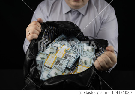 Having opened a large bag with money, the man shows it to the camera on a black background. A bag with packs of dollars in the hands of a businessman on a black background. 114125089