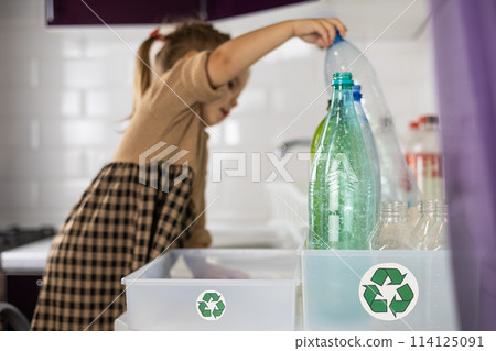 A girl carefully reaches across the kitchen table to place a plastic bottle in a trash box. Cute daughter in a dress helps her parents sort plastic for recycling. Zero waste concept. 114125091
